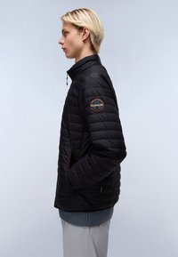 Zwarte gewatteerde jas met ritszakken, hoge kraag en logopatch op de mouw. Getextureerd oppervlak en getailleerde silhoette.