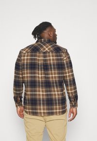 Camisa de franela a cuadros marrón y marino con mangas largas, puños con botones y dobladillo redondeado. La tela parece suave con un acabado texturizado.