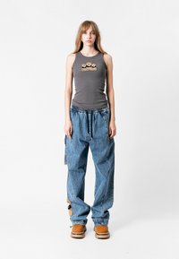 Top gris sin mangas con diseño bordado sobre el pecho, combinado con pantalones de mezclilla azules holgados con detalle de parche y cinturilla elástica.