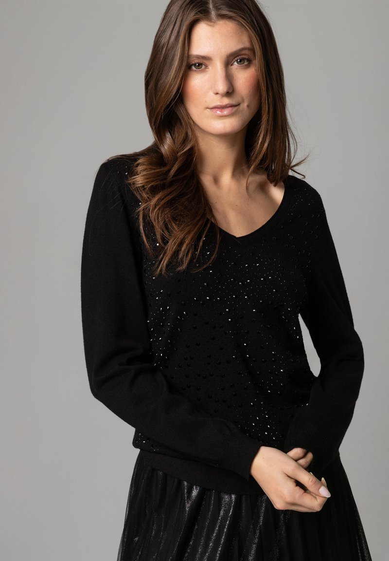 Schwarzer Strickpullover mit V-Ausschnitt, langen Ärmeln und strukturierten Details mit glänzenden Akzenten. Kombiniert mit einem plissierten schwarzen Rock.