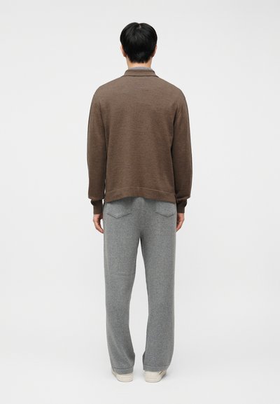 Άνδρας που στέκεται πλάτη, φοράει καφέ μακρυμάνικη μπλούζα, γκρι παντελόνι με πίσω τσέπες και λευκά αθλητικά παπούτσια μπροστά σε απλό φόντο.