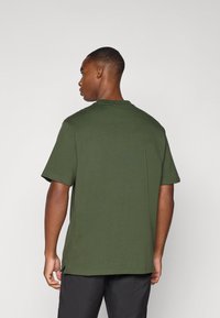 Olivenfarget bomull t-skjorte med korte ermer, rund hals og rett kant. Har en liten tagg på baksiden. Bæres med sorte bukser.