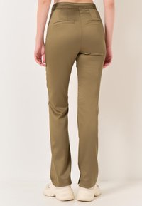 Khakifarbene Stretch-Hosen im Slim-Fit-Schnitt mit zwei Gesäßtaschen und einer glatten Textur, kombiniert mit beigen Sneakers vor einem neutralen Hintergrund.