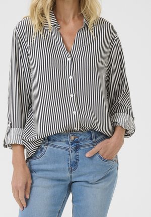 Frau trägt ein schwarz-weiß gestreiftes Hemd mit vertikalen Knöpfen, hochgekrempelten Ärmeln und hellblauen Jeans, eine Hand in der Tasche.