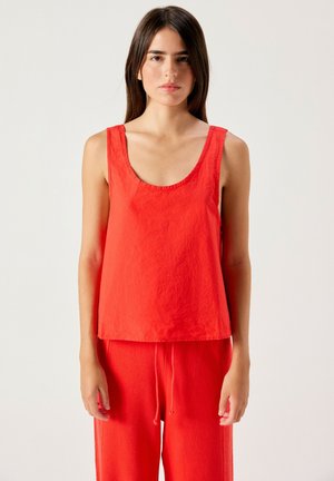 Giovane donna con capelli lisci e scuri che indossa un top rosso senza maniche e pantaloni rossi con coulisse, in piedi davanti a uno sfondo chiaro e uniforme.