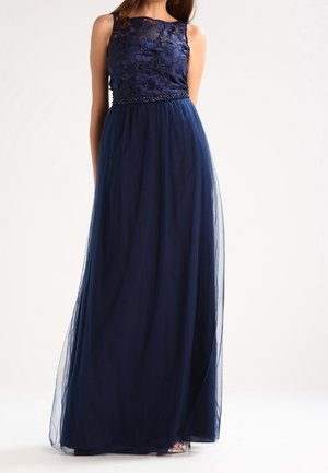Vrouw draagt een mouwloze marineblauwe avondjurk met een bloemrijke kanten lijfje en een vloeiende chiffon rok, staand tegen een effen achtergrond.