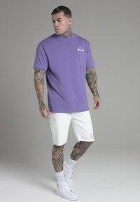 Lilla bomulds T-shirt med rund hals og korte ærmer, kombineret med hvide slidte shorts og hvide sneakers.