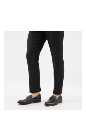 Beine in schwarzen, maßgeschneiderten Hosen und schwarzen Leder-Loafers mit Metall-Schnallendetail vor weißem Hintergrund.