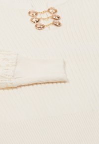 Witte geribbelde trui met een ronde halslijn, ingewikkelde kantafwerking op de mouwen en een decoratieve gouden ketting met versierde knopen.