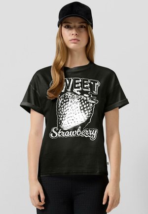 Jeune femme portant une casquette noire et un t-shirt noir avec un motif de fraise blanche et le texte "Sweet Strawberry", debout devant un fond gris clair.