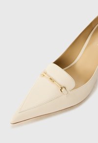Pointed-toe cream leather pump featuring a gold-tone hardware accent across the vamp. Smooth texture with stitched detailing.
