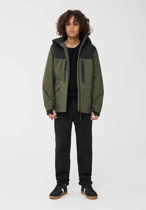 Giacca cappuccio verde e nera con tasche a zip, abbinata a pantaloni neri e sneakers. Presenta polsini elasticizzati e orlo regolabile.