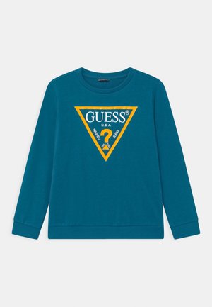 Bluza z długim rękawem w kolorze zielonkawoniebieskim z żółtym trójkątnym logo na środku z napisem „GUESS U.S.A. Washed Jeans” i znakiem zapytania w środku.