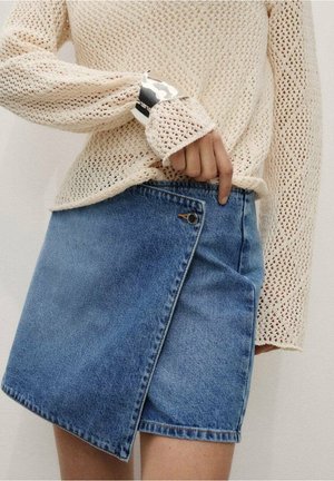 Persoon draagt een crème opengebreide trui en een blauwe denim wikkelrok met een asymmetrische voorflap en knoopsluiting.