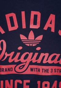 Černé tričko s výrazným červeným nápisem: "ADIDAS ORIGINALS" a ikonickým logem se třemi pruhy. Materiál působí měkce a hladce.