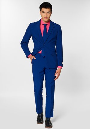 Young man stands confidently in a blue suit with a pink shirt and tie, hands on hips; black dress shoes complete the sharp look.
