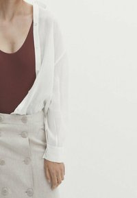 Witte linnen overhemd met knopen over een bordeaux bodysuit, gecombineerd met een beige rok met grote knopen en een subtiele textuur.