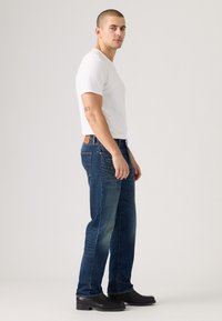 Jeans di denim blu con design a taglio dritto, caratterizzati da una toppa in pelle marrone chiaro sulla cintura, indossati con una maglietta bianca aderente e stivali neri.