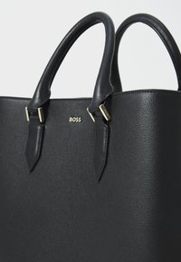 Borsa a tracolla in pelle nera con texture liscia, due manici superiori e dettagli in hardware dorato. Presenta un logo impresso sul fronte.