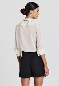 Camicia a criniera color crema chiaro con colletto arricciato e piping nero, abbinata a pantaloni corti neri a vita alta. Il tessuto ha una consistenza morbida.