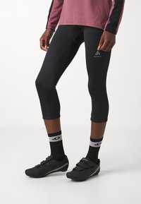 Fekete sportleggings, testhez simuló fazonnal, fekete kerékpáros cipőkkel, amelyek két pánttal rendelkeznek, valamint mintás zoknikkal, amelyeken egy logó látható.