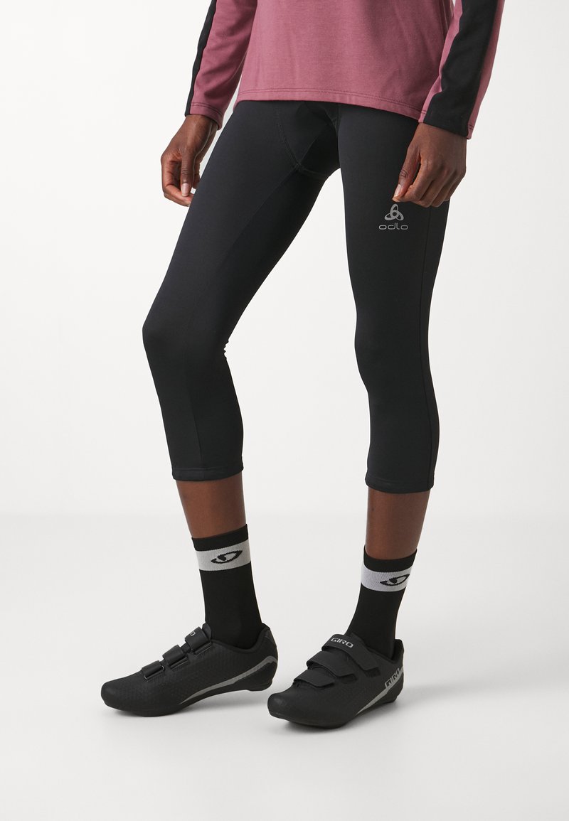 Fekete sportleggings, testhez simuló fazonnal, fekete kerékpáros cipőkkel, amelyek két pánttal rendelkeznek, valamint mintás zoknikkal, amelyeken egy logó látható.