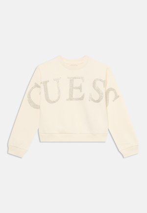 Sweat-shirt à manches longues couleur crème avec de grandes lettres en strass formant le mot "GUESS" sur le devant et les manches.