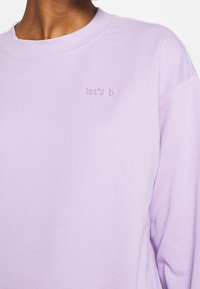 Sudadera color lilac hecha de tela suave, con cuello redondo y texto bordado "let's b" en el pecho. Diseño holgado.