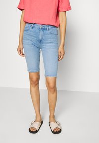 Bermuda shorts in denim azzurro chiaro con una silhouette aderente e orlo risvoltato, abbinati a una camicia a maniche corte color corallo e sandali beige.