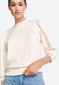 Pulover de culoare crem cu mâneci bufante, decolteu rotund și textură moale, asortat cu denim deschis. Finisaj neted și croială lejeră.