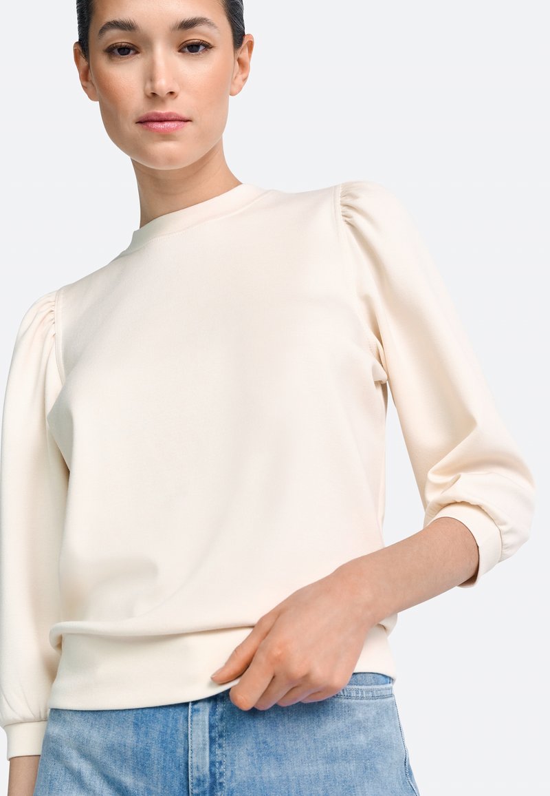 Pulover de culoare crem cu mâneci bufante, decolteu rotund și textură moale, asortat cu denim deschis. Finisaj neted și croială lejeră.