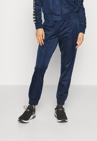 Partie inférieure du corps d'une personne portant un pantalon de survêtement bleu marine, une veste assortie avec des manches à motifs et des chaussures de sport noires.