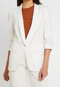 Blazer blanc à rayures avec un col cranté, fermeture à un bouton et manches retroussées. Associé avec un haut marron et un pantalon crème.