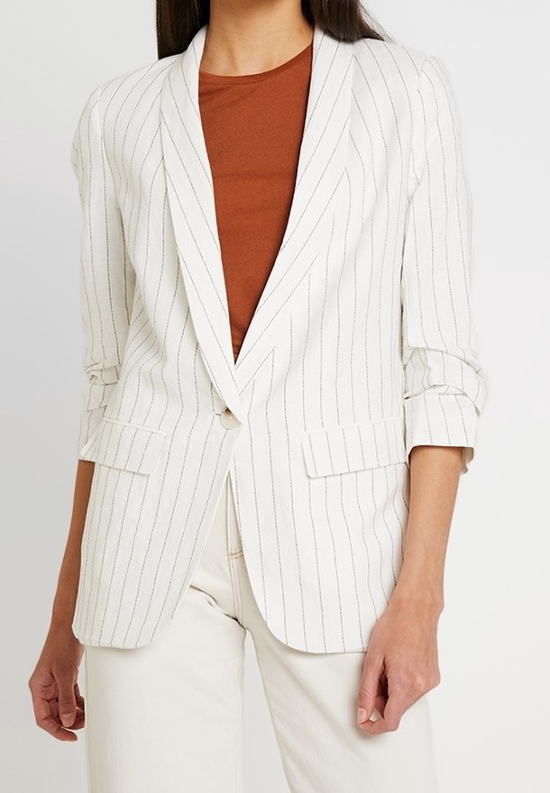 Blazer blanc à rayures avec un col cranté, fermeture à un bouton et manches retroussées. Associé avec un haut marron et un pantalon crème.