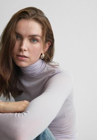 Haut col roulé violet clair à manches longues, en matière semi-transparente. Le mannequin a les cheveux ébouriffés et porte des boucles d'oreilles argentées en forme d'anneau.