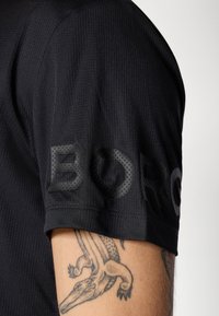Juodas trumpas rankovėmis marškinėlis su tekstūruotais "BORG" užrašais ant rankovės, rodančiais alkūnę su tatuiruote, vaizduojančia aligatorius arba krokodilą.