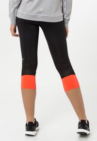Leggings negros con un diseño hasta la mitad de la pantorrilla y acentos en color naranja brillante en la parte inferior, combinados con una camiseta de manga larga gris y zapatillas de correr.