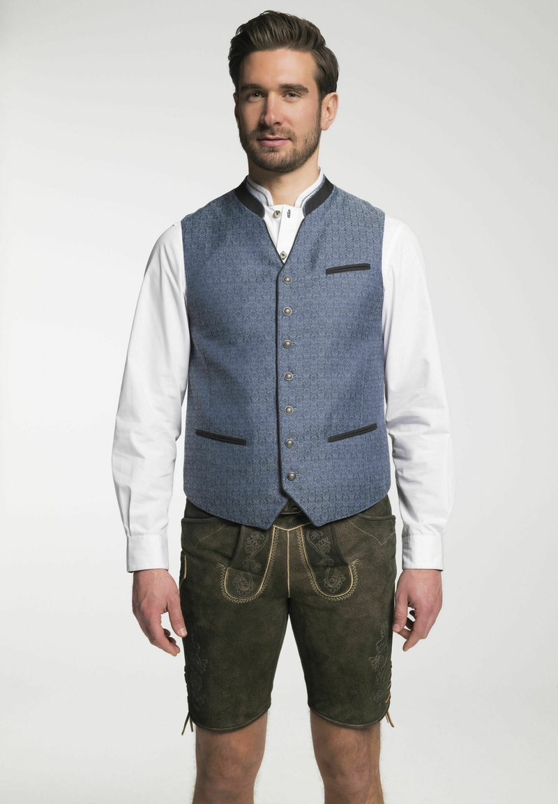 Traditionelles bayerisches Outfit mit einer blauen gemusterten Weste mit schwarzen Akzenten und einem weißen Hemd, kombiniert mit dunklen braunen Lederhosen mit dekorativen Nähten.