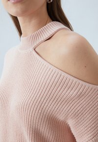 Pull tricoté rose clair avec un col montant côtelé et un design à épaules dénudées. La texture est douce et légèrement texturée.