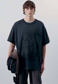 Camisa de algodón negra de corte oversized con un gran diseño gráfico tonal. La modelo sostiene un bolso marrón. Ajuste simple y relajado con mangas cortas.