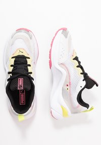 Chaussures de sport blanches avec une tige en mesh, des lacets noirs et des accents roses sur la semelle. Panneaux texturés avec des détails jaunes et gris.