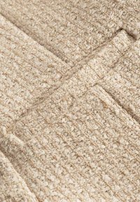 Gros plan d’un textile tissé beige avec un motif texturé bouclé et des coutures cousues créant des formes géométriques.