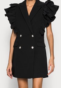 Svart ärmlös blazerklänning med volangaxlar, dubbelradiga kristallknappar och fickor med klaff bärs av en person.