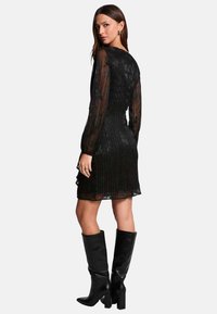 Robe noire plissée avec des manches longues en organza, une silhouette ajustée et un ourlet en dents de scie. Associée à des bottes en cuir noir jusqu&#x27;aux genoux.