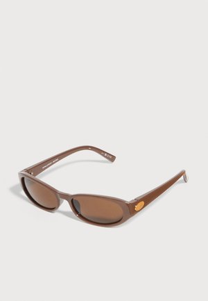 Lunettes de soleil rectangulaires marron avec des verres légèrement courbés et des branches fines, posées sur une surface gris clair.