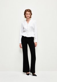 Blusa bianca a peplo con scollatura a V profonda e chiusura con bottoni frontali, abbinata a pantaloni neri a palazzo e tacchi neri. Tessuto liscio e design strutturato.