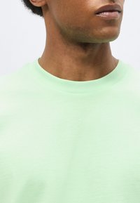 Κοντινό πλάνο ενός ατόμου που φοράει μια ανοιχτοπράσινη μπλούζα με στρογγυλή λαιμόκοψη, δείχνοντας τον λαιμό, τη γνάθο και μέρη των χειλιών σε απλό φόντο.