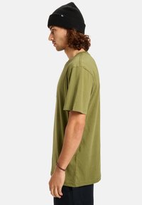 Olivově zelené bavlněné tričko s krátkým rukávem, kulatým výstřihem a uvolněným střihem. Nosené s černou pletenou čepicí.