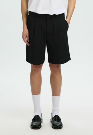 Homme portant un short noir plissé, des chaussettes blanches, des mocassins noirs et une chemise blanche, debout les mains dans les poches sur fond blanc.