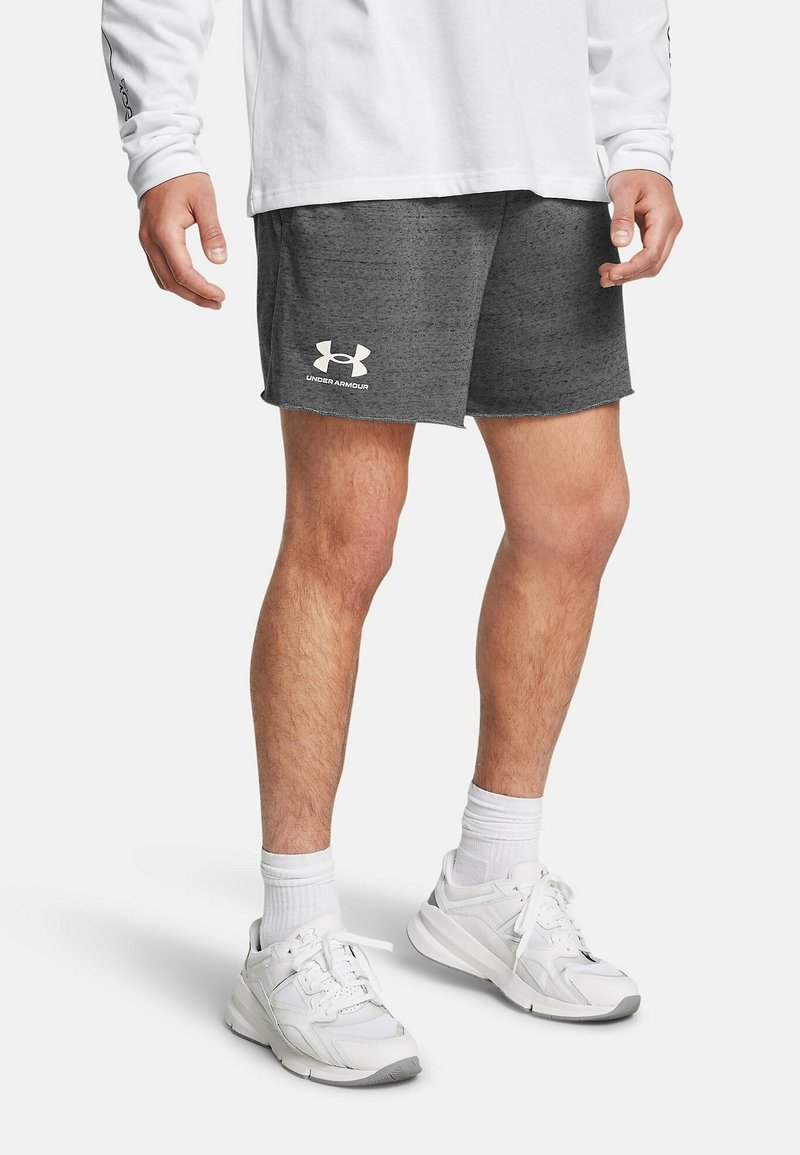 Homme portant un short gris Under Armour, un t-shirt blanc à manches longues, des chaussettes blanches hautes et des chaussures de sport blanches, debout sur un fond uni.